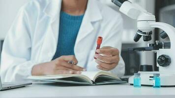 Medical worker in lab coat and sterile mask, doing a microscope analysis while her colleague are working behind video
