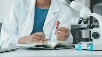 Medical worker in lab coat and sterile mask, doing a microscope analysis while her colleague are working behind video