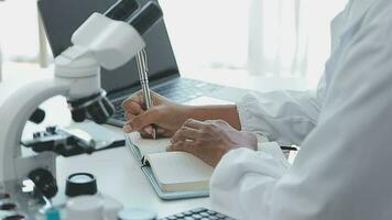 Medical worker in lab coat and sterile mask, doing a microscope analysis while her colleague are working behind video