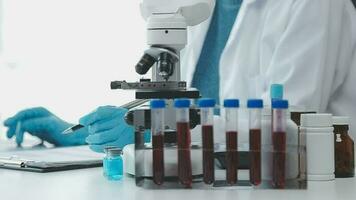 Medical worker in lab coat and sterile mask, doing a microscope analysis while her colleague are working behind video