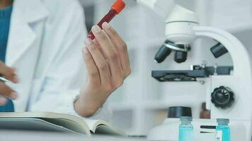 Medical worker in lab coat and sterile mask, doing a microscope analysis while her colleague are working behind video