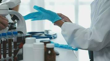 Medical worker in lab coat and sterile mask, doing a microscope analysis while her colleague are working behind video