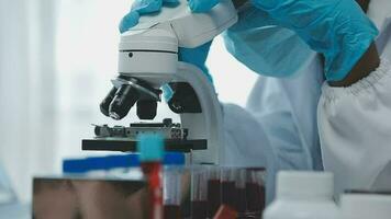 Medical worker in lab coat and sterile mask, doing a microscope analysis while her colleague are working behind video