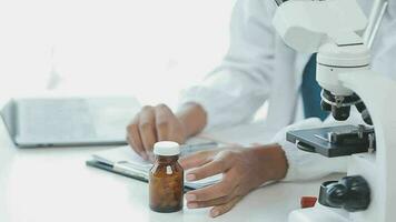 médico trabajador en laboratorio Saco y estéril mascarilla, haciendo un microscopio análisis mientras su colega son trabajando detrás video