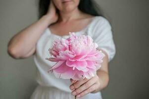 A young woman in a white dress holds a pink peony in her hands. photo