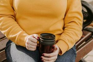 Young red-haired woman holds reusable mug in her hands. She sits on park bench and is wearing yellow sweater. Lifestyle on sunny autumn day. photo