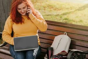 Young red-haired woman freelancer works in park using laptop. Modern businesswoman talking on phone. Online work on Internet. photo