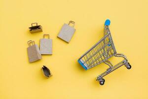 Eco-friendly craft paper bags next to a metal shopping cart on a yellow background. Black Friday, gift sales. Top view photo