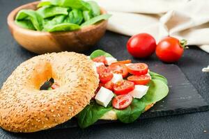 Appetizing bagels stuffed with tomatoes, feta and spinach leaves on a slate board on a black background. Light healthy snack. Close-up photo