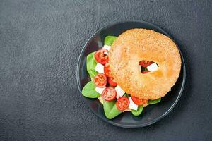 Appetizing ready-to-eat bagels stuffed with tomatoes, feta and spinach leaves on a plate on a black background. Light healthy snack. Top view. Copy space photo