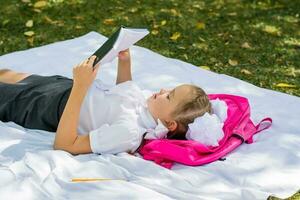 A young schoolgirl is doing homework lying on a blanket and a backpack in a sunny autumn park. Outdoor education for children. Back to school concept photo