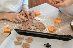 preparando a celebrar Víspera de Todos los Santos y preparando un tratar. niños cortar fuera horneando sartenes para Víspera de Todos los Santos galletas y un horneando sábana de crudo galletas en el mesa. estilo de vida foto