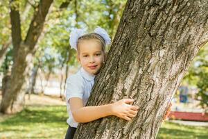 un linda Chica de escuela con blanco arcos soportes por un árbol en un soleado otoño parque. primero grado en septiembre 1er. espalda a colegio concepto foto