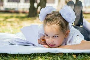 de cerca retrato de un pequeño Chica de escuela haciendo deberes mientras acostado en un cobija en un soleado otoño parque. al aire libre educación para niños. espalda a colegio concepto foto