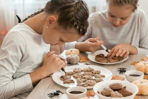 Two girls decorate halloween gingerbread cookies on plates with chocolate icing. Cooking treats for halloween celebration. Lifestyle photo