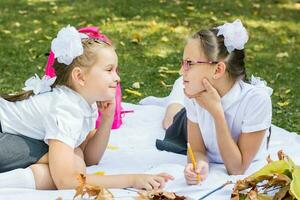 dos linda colegialas son sonriente y haciendo deberes en un cobija en un soleado otoño parque. al aire libre educación. espalda a colegio concepto foto