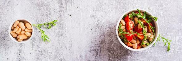 Homemade salad of white beans, cherry tomatoes and arugula in a bowl top view web banner photo