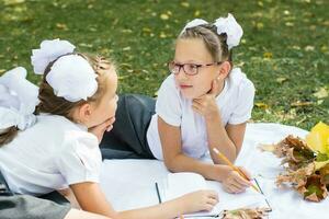 dos linda colegialas Mira a cada otro, sonrisa y reflejar en deberes en un cobija en un soleado otoño parque. al aire libre educación. espalda a colegio concepto foto