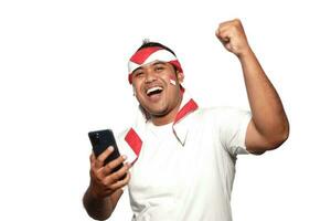 emocionado asiático hombre vistiendo blanco camiseta sonriente mientras participación su teléfono, aislado por blanco antecedentes. indonesio independencia día celebracion foto