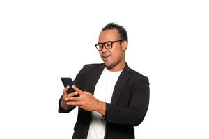 emocionado asiático hombre con lentes vistiendo negro traje sonriente mientras participación su teléfono, aislado por blanco antecedentes foto