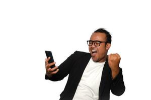 Excited Asian man with glasses wearing black suit smiling while holding his phone, isolated by white background photo