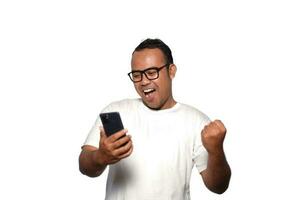 Excited Asian man with glasses wearing white Tshirt smiling while holding his phone, isolated by white background photo