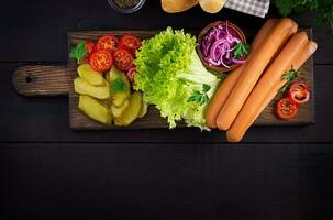 Ingredients for hot dog with  sausage, pickled cucumber, tomatoes, red onions and lettuce on dark wooden background. Summer hotdog. Top view photo