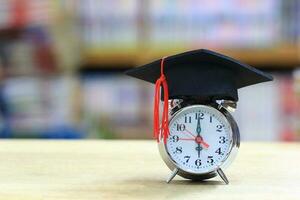 Alarm clock with graduation hat on bookshelf in the library room background, Saving money for education concept photo