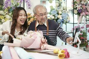 E-commerce business, Asian elder man and daughter florist shows floral arrangements and talks to camera via online live-streaming webcam application in a bright flower shop, beautiful blossoms store. photo