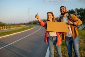 Happy couple hitchhiking on roadside trying to stop car. They are holding blank cardboard for your text. photo