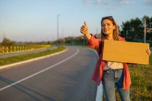 Woman is hitchhiking on roadside trying to stop car. She is holding blank cardboard for your text. photo