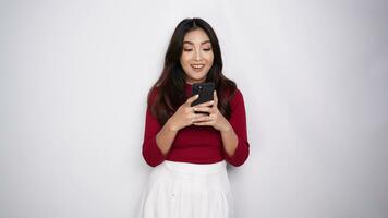 un retrato de un contento asiático mujer vistiendo un rojo camisa mientras participación su teléfono, aislado por blanco antecedentes video