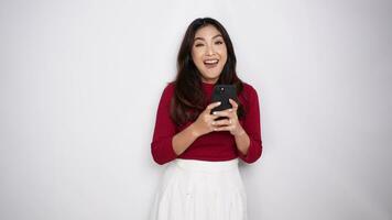 uma retrato do uma feliz ásia mulher vestindo uma vermelho camisa enquanto segurando dela telefone, isolado de branco fundo video