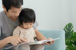 Happy Asian father relax and read book with baby time together at home. parent sit on sofa with daughter and reading a story. learn development, childcare, laughing, education, storytelling, practice. photo