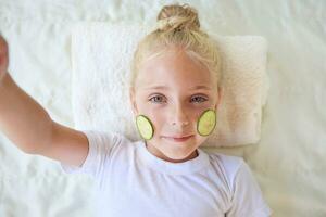 Beautiful girl with facial mask of cucumber taking selfie while posing to phone. photo