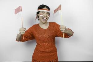 Happy smiling Indonesian woman wearing red kebaya and headband holding Indonesia's flag to celebrate Indonesia Independence Day isolated over white background. photo