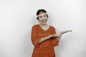 Excited Asian woman wearing red kebaya and headband, pointing at the copy space beside her, isolated by white background. Indonesia's independence day concept. photo