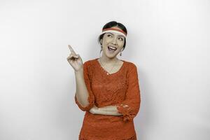 Excited Asian woman wearing red kebaya and flag headband, pointing at the copy space on top of her, isolated by white background photo