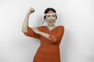 Excited Asian woman wearing a red kebaya and headband showing strong gesture by lifting her arms and muscles smiling proudly. Indonesia's independence day concept. photo