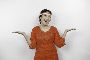 Excited Asian woman wearing red kebaya and headband, pointing at the copy space beside her, isolated by white background. Indonesia's independence day concept. photo