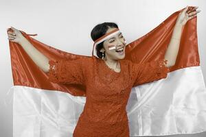 Happy smiling Indonesian woman wearing red kebaya and headband holding Indonesia's flag to celebrate Indonesia Independence Day isolated over white background. photo