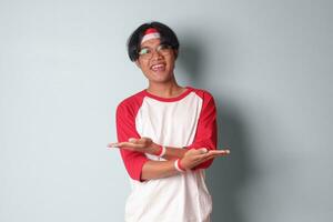 Portrait of attractive Asian man in t-shirt with red and white ribbon on head, choosing between 2 different options, holding two hands with empty space. Decision concept. Isolated image on gray photo