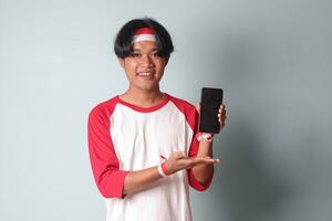 Portrait of attractive Asian man in t-shirt with red and white ribbon on head, holding and showing blank screen of mobile phone for mock-up. Isolated image on gray background photo