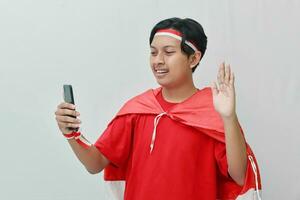 retrato de atractivo asiático hombre en camiseta con rojo blanco cinta en cabeza con bandera en su hombro como un capa, ondulación mano diciendo Hola en su móvil teléfono durante vídeo vocación. aislado imagen en gris foto