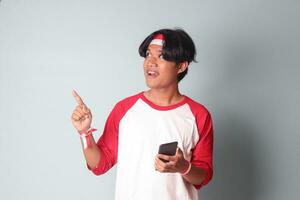 Portrait of attractive Asian man in t-shirt with red and white ribbon on head, holding mobile phone while raising up Indonesia flag and pointing away. Isolated image on gray background photo