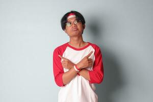 Portrait of attractive Asian man in t-shirt with red and white ribbon on head, choosing between 2 different options, holding two hands with empty space. Decision concept. Isolated image on gray photo