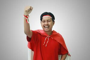 Portrait of attractive Asian man in t-shirt with red white ribbon on head with flag on his shoulder as a cloak, raising his fist, celebrating Indonesia's independence day. Isolated image on gray photo