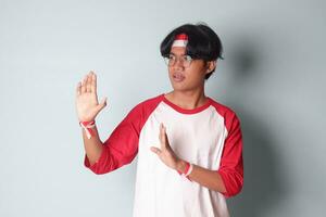 Portrait of attractive Asian man in t-shirt with red and white ribbon on head, forming a hand gesture to avoid something. Advertising concept. Isolated image on gray background photo