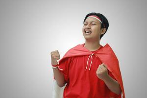 Portrait of attractive Asian man in t-shirt with red white ribbon on head with flag on his shoulder as a cloak, raising his fist, celebrating Indonesia's independence day. Isolated image on gray photo