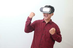 Portrait of Asian man in red plaid shirt using Virtual Reality VR glasses and raising his fist, celebrating success. Isolated image with copy space on white background. photo